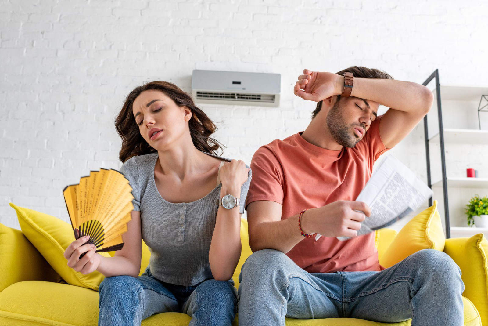 A man and woman sitting on a couch feeling uncomfortably hot indoors while trying to cool down with a handheld fan.